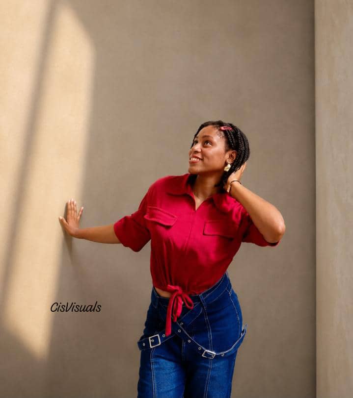 Woman in a red shirt and belted denim leaning against a sunlit tan wall